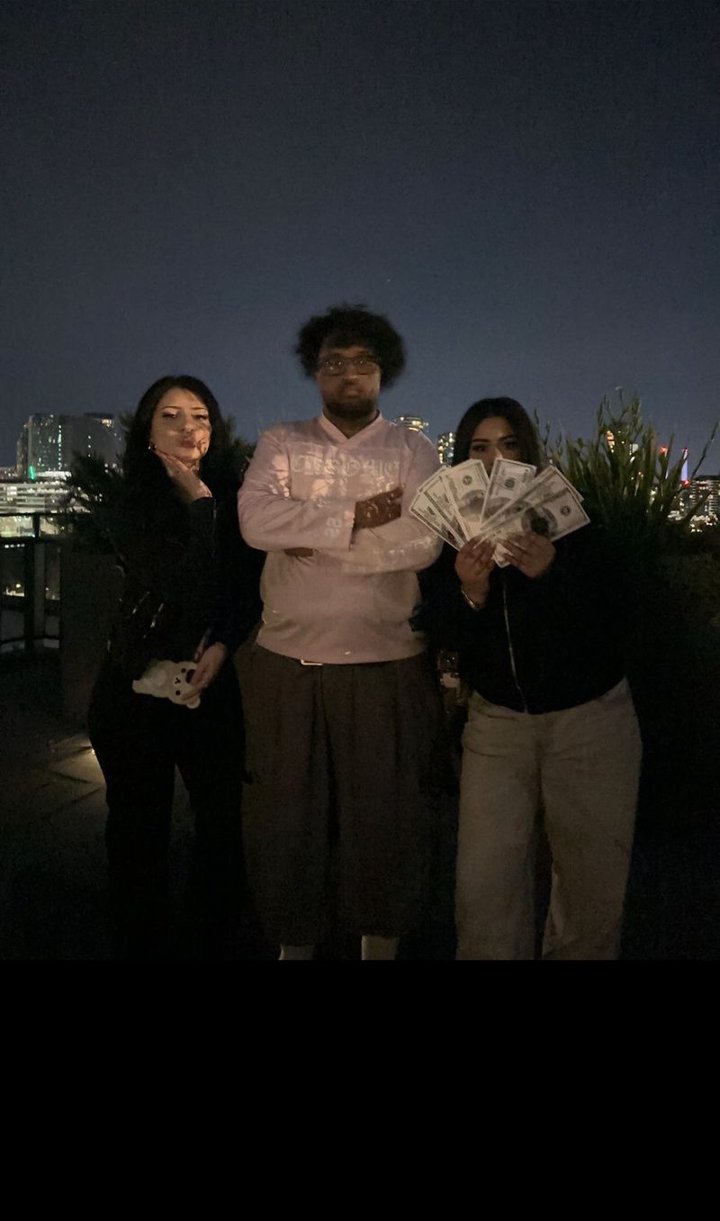 Three friends on a rooftop at night with a city skyline behind them, one holding a fanned stack of Mahdi Bucks